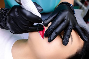 Kalıcı bir makyöz bir modelin önünde oturur ve dudak dövmesi yaptırır. Dövme makinesi ve kırmızı dudak pigmentiyle.