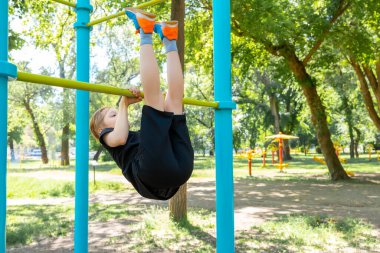 Sporcu çocuk yatay barda jimnastik yapıyor, bacaklarını kaldırıyor ve kendini yukarı çekiyor.