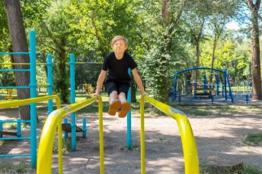 Sporcu çocuk, jimnastik çubuklarında egzersiz yapıyor. Bacaklarını ileri uzatıyordu ve bu egzersizi yapmanın onun için çok zor olduğu gerçeğinden yüzünü büküyordu.