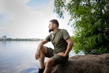 Sakallı bir adam kayaya oturur ve düşünceli bir şekilde nehre bakar.