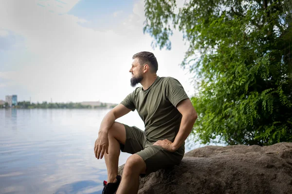 Sakallı bir adam kayaya oturur ve düşünceli bir şekilde nehre bakar.