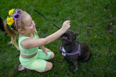 little cheerful girl in a green suit next to a french bulldog breed dog sitting on the lawn in the park