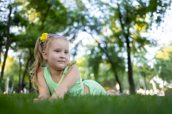 a little cheerful girl in a green suit lies on the grass in the park and looks away