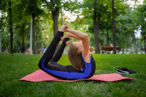 in the park, a little girl bends back, clutching her legs, performs yoga elements, does stretching
