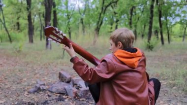 Çocuk arkası dönük oturur ve sönmüş bir ateşin önünde gitar çalar.