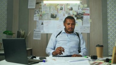 Sigara içen dedektif masada oturuyor, sandalyeye dönüyor ve soruşturma kuruluna bakıyor. Polis memuru ya da FBI ajanı. Soruşturma kurulu ofisinde kılıflı. Yüksek kalite 4K görüntü.