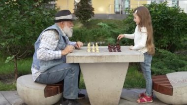 Zoom in grandpa playing chess with beloved granddaughter outdoor. Multi-generational family happily spending time together engaged in educational board game. Happy multi-generational family concept