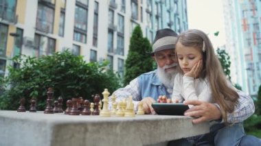 Lovely granddaughter teaches grandpa to use mobile phone outdoor. Cute little girl playing on smartphone with grandfather instead of chess. Grandparent learn using modern technology gadget concept.