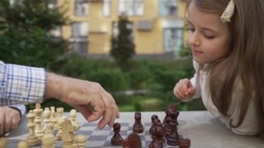 Grandpa teaches smart grandchild to play chess outdoor. Multi-generational family engaged in educational board game. Kid development, gain abstract thinking skills, hobby concept.