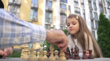 Close up smart concentrated small girl plays chess with grandfather outdoor. Brilliant little girl playing chess with her senior master. Kid development, gain abstract thinking skills, hobby concept.