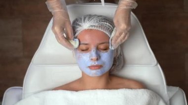 Güzellik uzmanının kadın müşteri yüzünden mavi maskeyi çıkarması. Genç bir bayan yüz bakımı yaptırıyor. Güzellik kliniği, kaplıca ve cilt bakımı konsepti. Yüksek kalite 4k görüntü