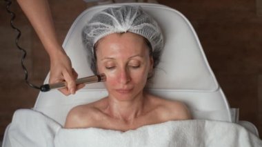 Güzel, 40 'lı yaşlarda orta yaşlı bir kadının kozmetik kliniğinde cildini gençleştirmek için donanım kozmetolojisi kullandığı bir fotoğraf. Cerrahi olmayan, donanım kozmetolojisi, gençleşme karşıtı cilt bakımı konsepti.