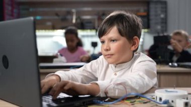 Bilgisayarı kullanarak robotik mühendisliği sınıfı için programlanan, odaklanmış zeki beyaz öğrenci portresi, kameraya bakmak ve arka planda okul sınıfıyla gülümsemek. STEM eğitim konsepti 4K