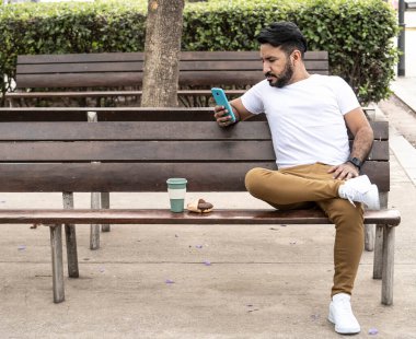 Plaza bankında oturan Latin adam elinde kahve ve çörekle cep telefonuna bakıyor.
