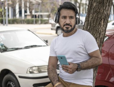 Şehirdeki bir ağaca yaslanmış sakallı, cep telefonuyla müzik dinleyen Latin bir adam.