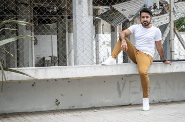 Arka plandaki inşaat alanında, alçak duvarda oturan sakallı bir Latin.