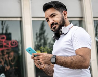 Sakallı Latin bir adam, cep telefonu kullanarak kameraya bakıyor.