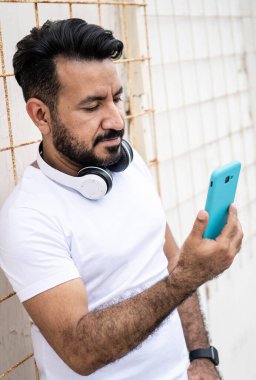 Sakallı Latin bir adam, cep telefonuna bakıp duvara yaslanıyor..