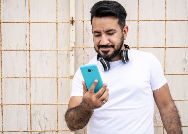 Sakallı Latin bir adam, cep telefonuna bakıp duvara yaslanıyor..