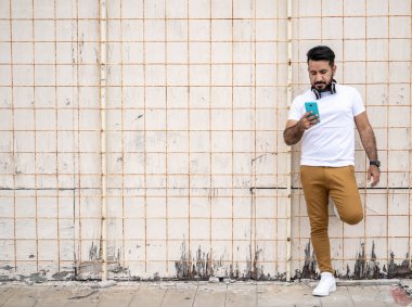 Sakallı Latin bir adam, cep telefonuna bakıp duvara yaslanıyor..