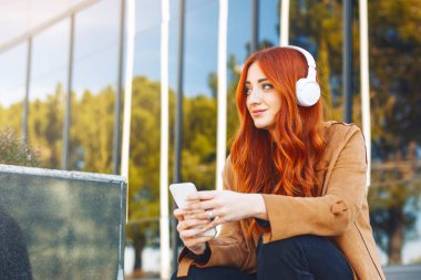 Kablosuz kulaklıkları ve akıllı telefonlarıyla müzik dinleyen genç kızıl saçlı kadın.,