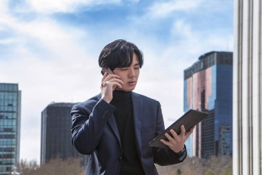 İş merkezinin dışında Asyalı bir adam var. Tabletine bakarken cep telefonuyla konuşuyor.
