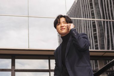 İş yerinin dışındaki Asyalı adam cep telefonuyla konuşuyor, cam binaların arka planı... 