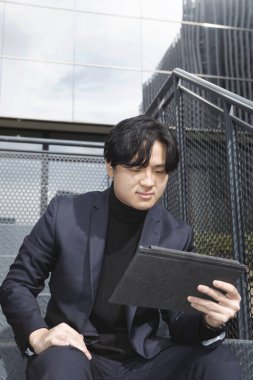 Asyalı bir adam merdivenlerde oturmuş iş merkezinin dışındaki masasına bakıyor.