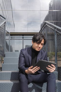 İş merkezinin dışında Asyalı bir adam merdivenlerde oturmuş tabletine bakıyor.