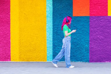 Pembe saçlı genç kız, renkli bir sokakta yürürken müzik dinliyor..