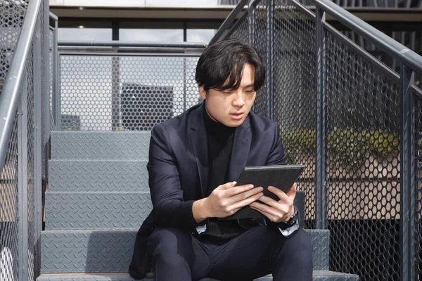 Asyalı bir adam merdivenlerde oturmuş tabletine endişeli bir şekilde bakıyor.
