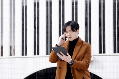 İş merkezinin dışında Asyalı bir adam tabletini kullanıyor ve cep telefonuyla konuşuyor. İş konsepti. Yüksek kalite fotoğraf