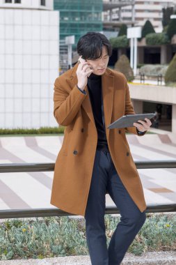 İş merkezinin dışında Asyalı bir adam tabletini kullanıyor ve cep telefonuyla konuşuyor. İş konsepti. Yüksek kalite fotoğraf