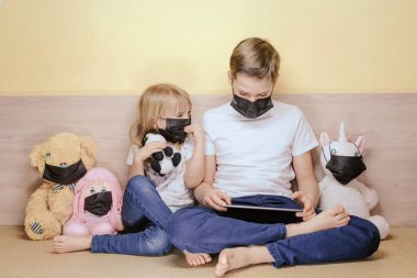 Erkek ve kız maskeler yüz maskesi takan oyuncaklar arasında tablet kullanıyorlar. COVID-19 koronavirüs salgını konsepti.