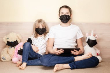 Erkek ve kız maskeler yüz maskesi takan oyuncaklar arasında tablet kullanıyorlar. COVID-19 koronavirüs salgını konsepti.