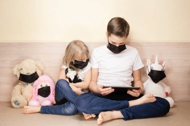 Erkek ve kız maskeler yüz maskesi takan oyuncaklar arasında tablet kullanıyorlar. COVID-19 koronavirüs salgını konsepti.