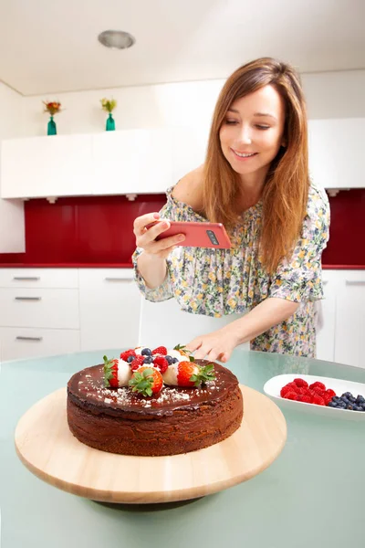 Genç sarışın beyaz kadın gülümsüyor ve cep telefonuyla sosyal medya için çikolatalı cheesecake 'inin fotoğraflarını çekiyor.