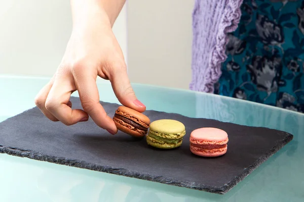 3 macarons, brown, green and pink, presented on a culinary blackboard. young woman's hand catching one of them. The art of pastry.
