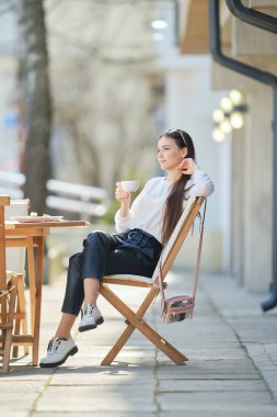 Güneşli bir günde kafenin terasında oturan ve kahve içen düşünceli genç bir kadın.. 