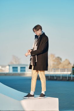 Eski yün ceketli, eşarplı ve kısa pantolonlu genç bir adam profili açık havada duruyor.