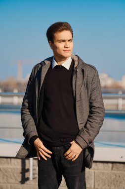 Gri ceketli genç adam, siyah kazak, beyaz gömlek ve sokakta poz veren siyah kot.