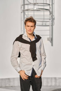 Beyaz gömlekli ve süveterli genç adam omuzlarından sarkıyor caddede yürüyor.