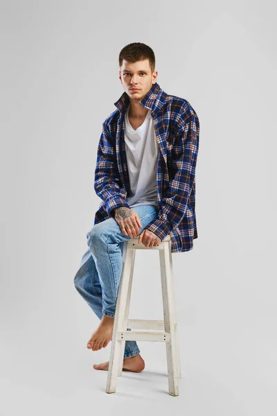 穿着衬衫和牛仔裤坐在高高的木椅上的年轻人的全长肖像 — 图库照片