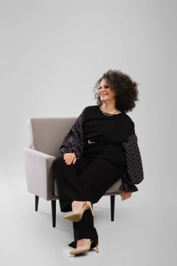 Kıvırcık saçlı mutlu bir son sınıf kadını gri arka planda, stüdyo koltuğunda oturuyor.