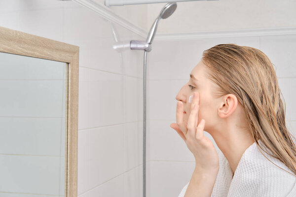 Young woman standing in profile and applying moisturizing cream on face  after shower. Skin care daily routine.