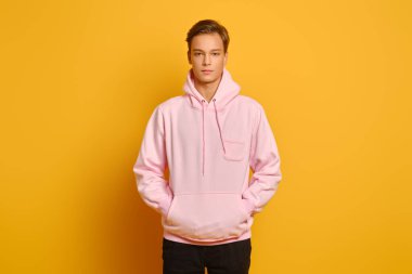 Pembe eşofmanlı düşünceli genç adam sarı stüdyo arka planında elleri cebinde poz veriyor.