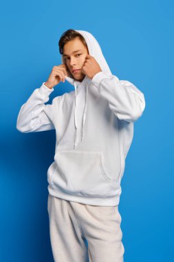 Beyaz eşofmanlı düşünceli genç adam mavi stüdyo arka planına kapüşon takıyor.
