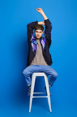 Rüzgarlık ceketli genç adam, zeytin gömlekli, kot pantolonlu ve çoraplı adam mavi stüdyo arka planında, uzun sandalyede oturuyor.