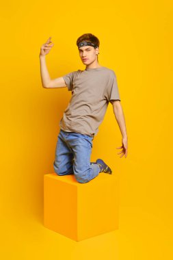 Genç adam dizlerinin üzerine çöküp, parlak sarı arka planda jest yapıyor.