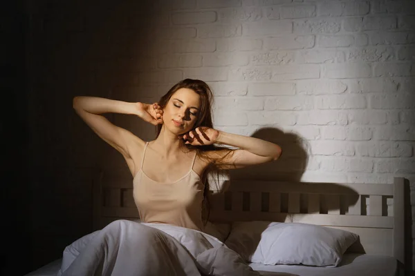 睡眠不足的漂亮女人早上醒来后躺在床上 把胳膊伸到黑暗的卧室的两侧 — 图库照片